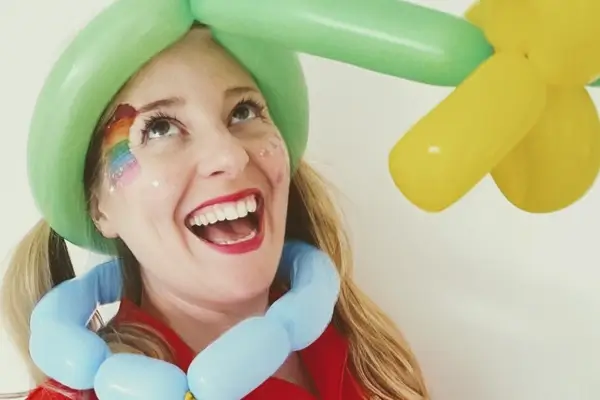 Corinne Wilkinson with a balloon flower hat and glitter rainbow face paint, looking up and grinning.